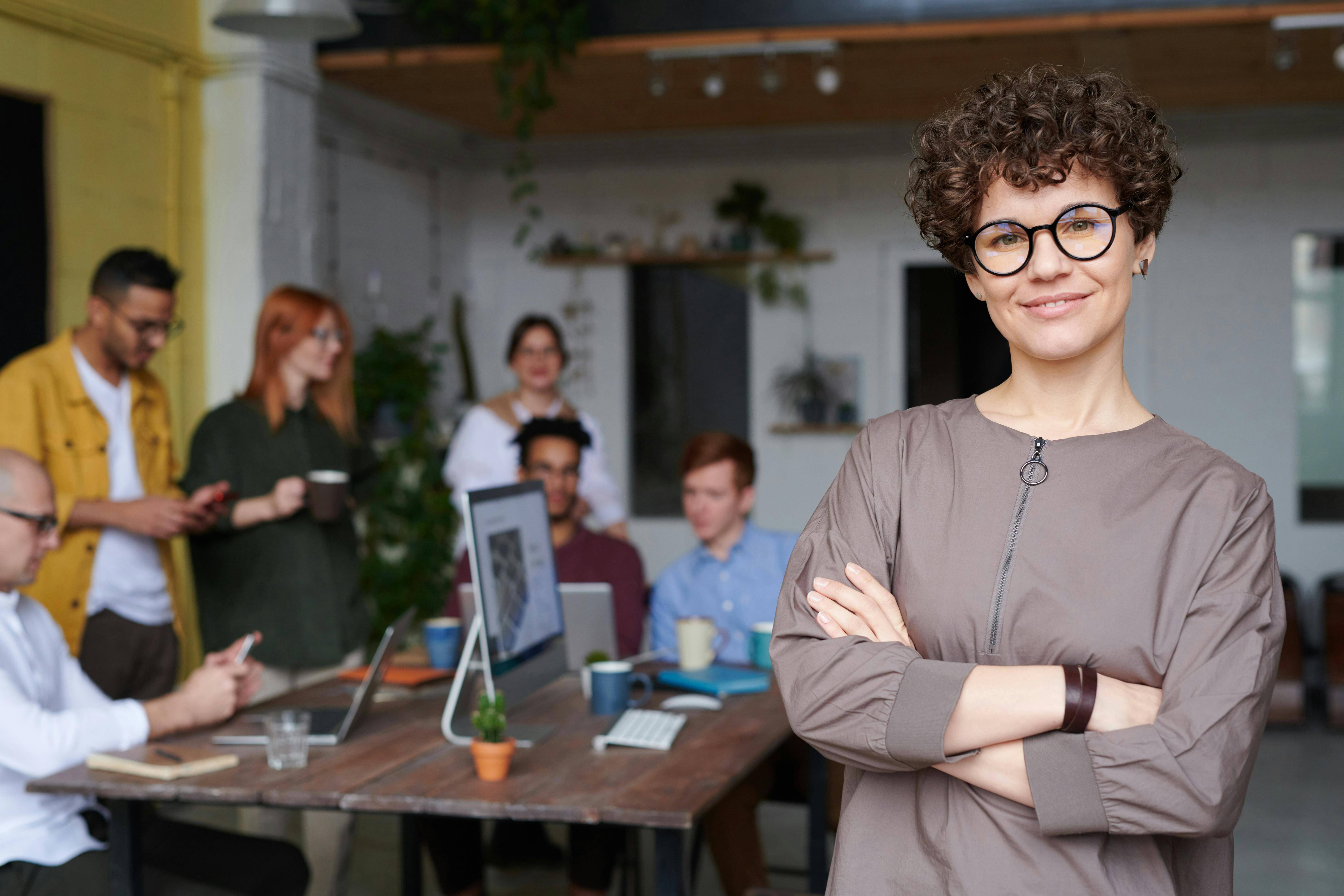 A woman wearing glasses stands confidently in front of a diverse group of people, engaging them with her presence.