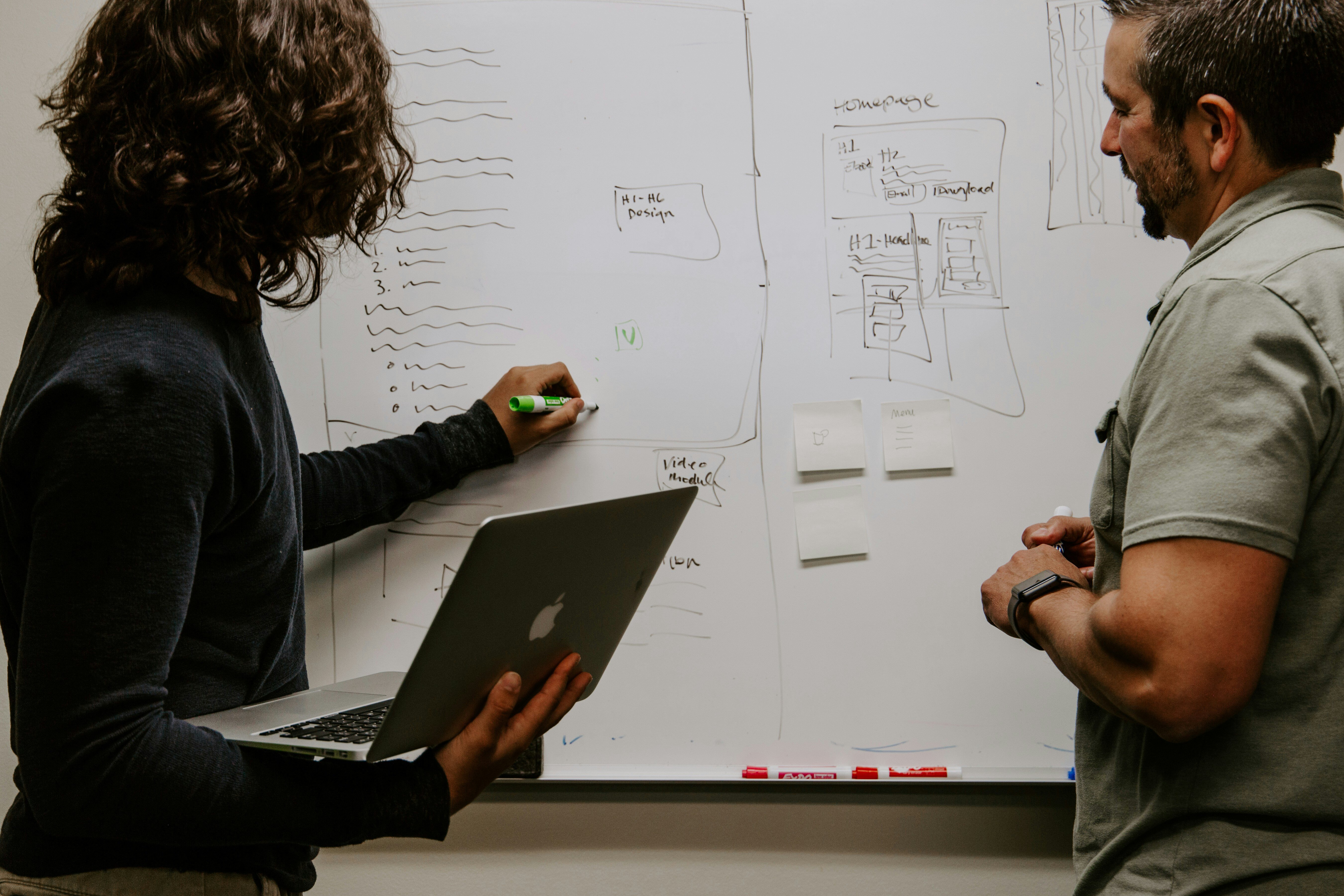 Two men standing in front of a whiteboard, engaged in discussion and pointing at various notes and diagrams.